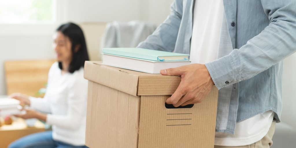 A man holds a moving box with the blurred image of a woman in the background.