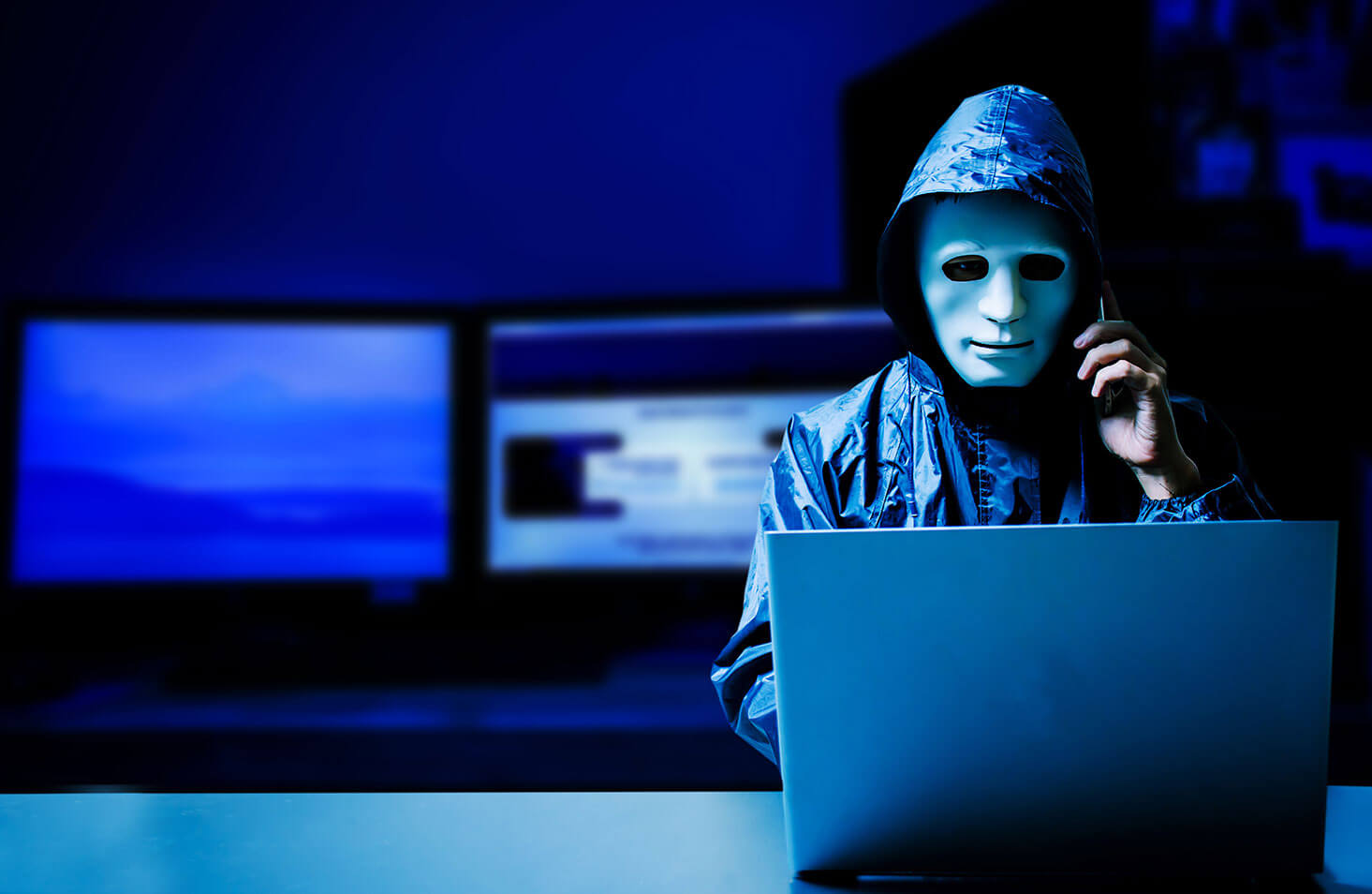 a man wearing a hooded jacket and with a sculpted mask over his face speaks on a phone while looking at a laptop in a dark room