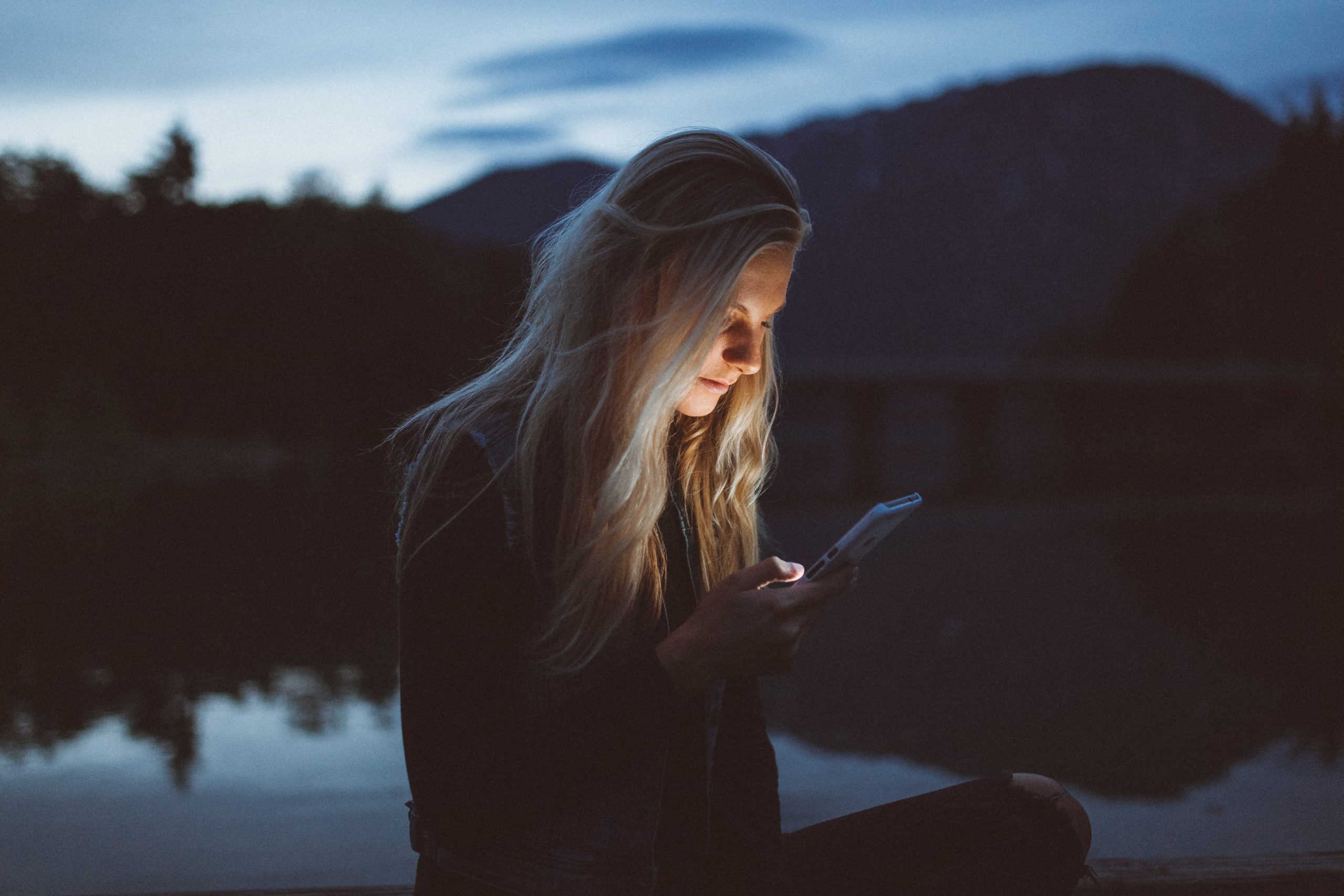 A young white woman with long blond hair looks at a phone, her face illuminated by its glow. She stands in a natural area with a lake and mountains. (Photo by Becca Tapert on Unsplash) 