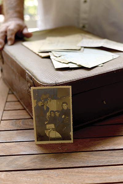 Photograph showing a suitcase, a few letters, and a photograph from the 1950s in the foregroundd, with a man's torso and hand in the background 