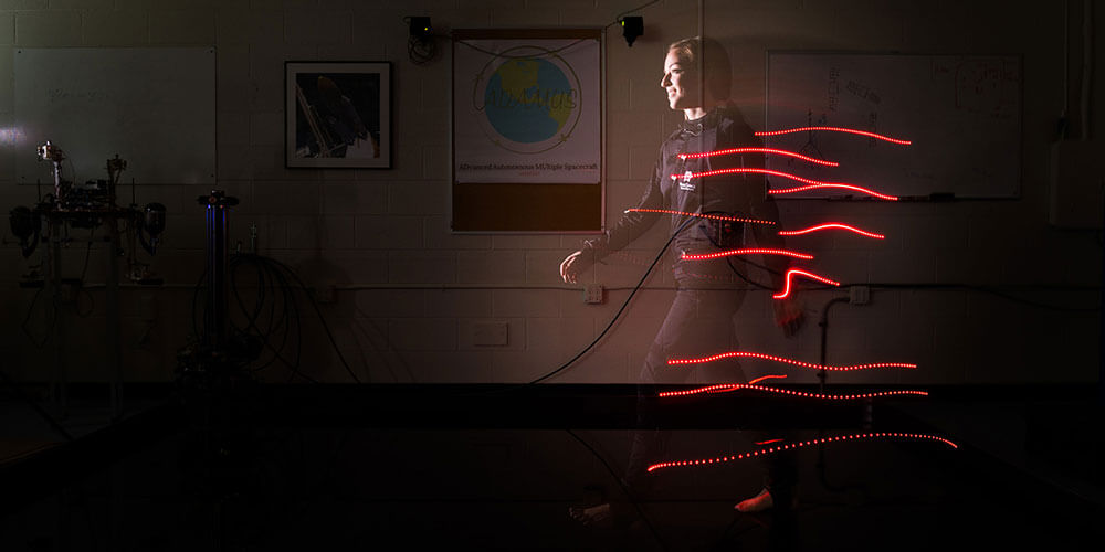 A semi-transparent projection of a smiling white woman walks through a dim classroom, trailing strings of red light behind her