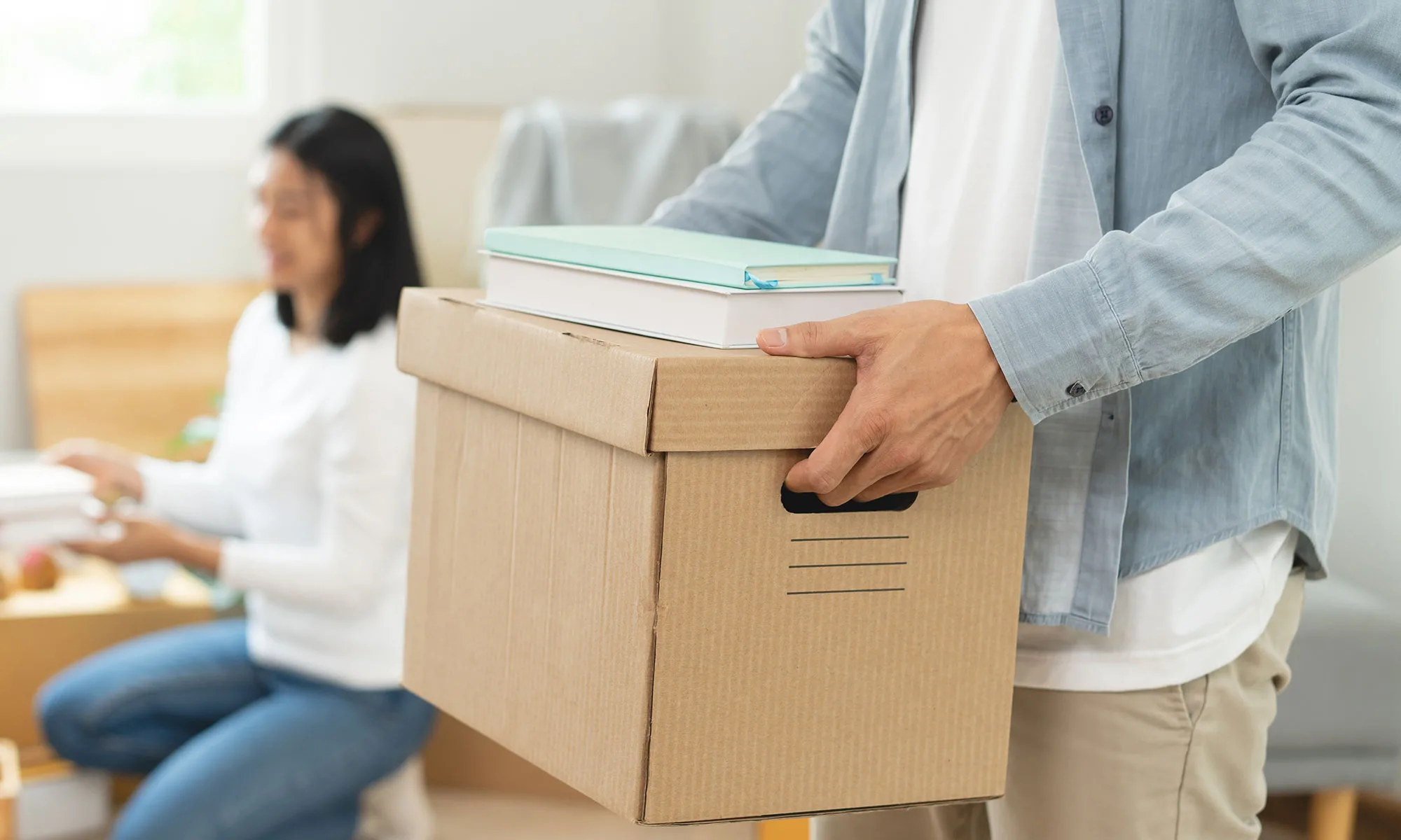A man holds a moving box with the blurred image of a woman in the background.