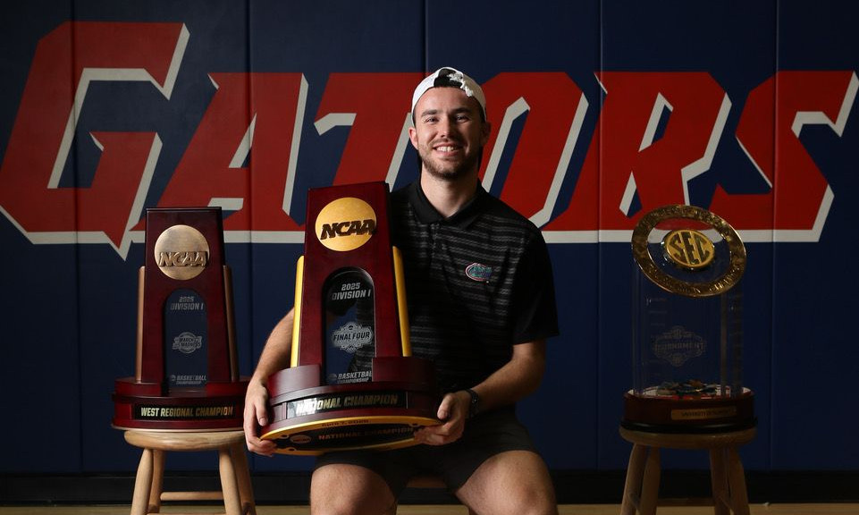 Corey Reynolds sitting in the basketball gym holding a championship trophy, with two additional trophies displayed beside him.