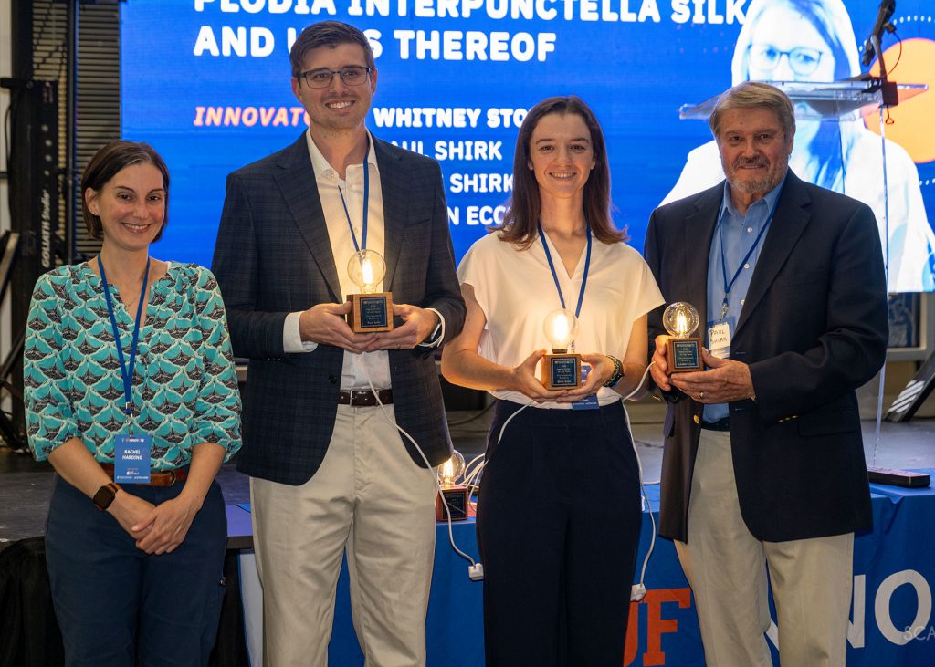 Graduate students in the Stoppel lab, Bryce Shirk and Lauren Eccles (center) accept an award on behalf of Whitney Stoppel, Ph.D. associate professor in Chemical Engineering for their project on engineering the Indianmeal moth to produce silk fibers containing therapeutic proteins. Paul Shirk from the USDA is on the right and Rachel Harding, the UF Innovate tech licensing officer, is on the left.