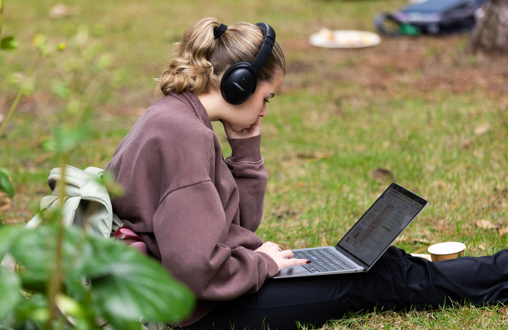 A student wearing headphones sits on the grass outside and works on a laptop.