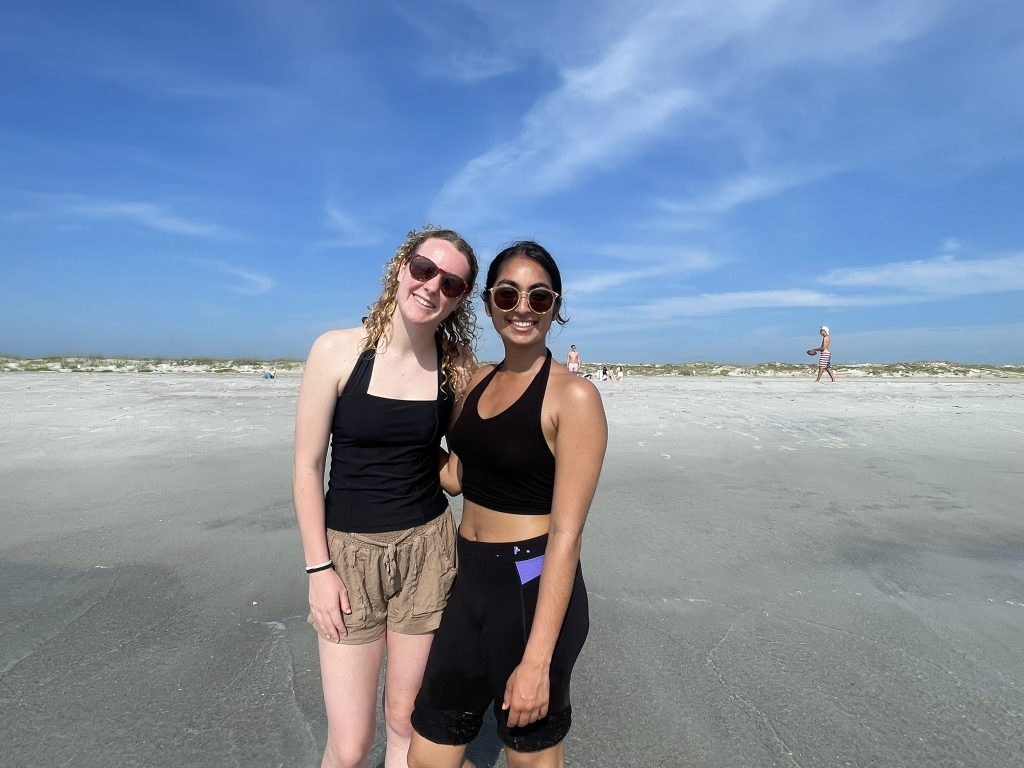 Two female students wearing sunglasses posing together on the shoreline. 