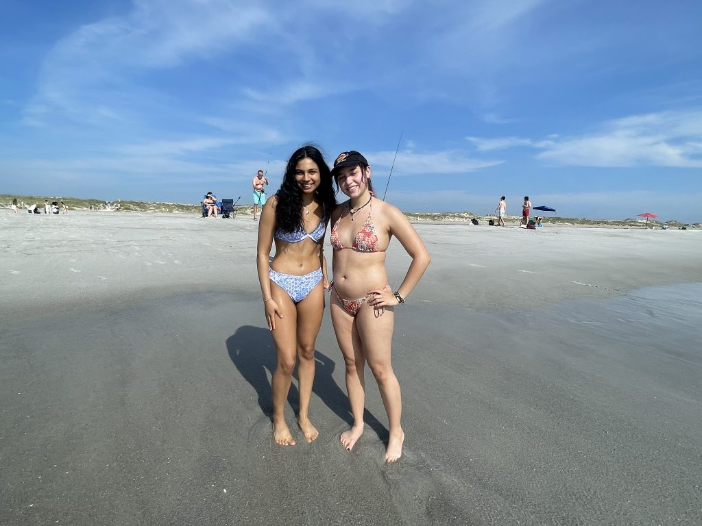 Two female students wearing bathing suits posing together on the shoreline. 