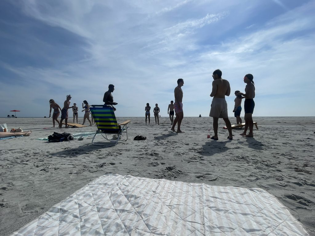 A group of students standing around and playing cornhole on the beach. 