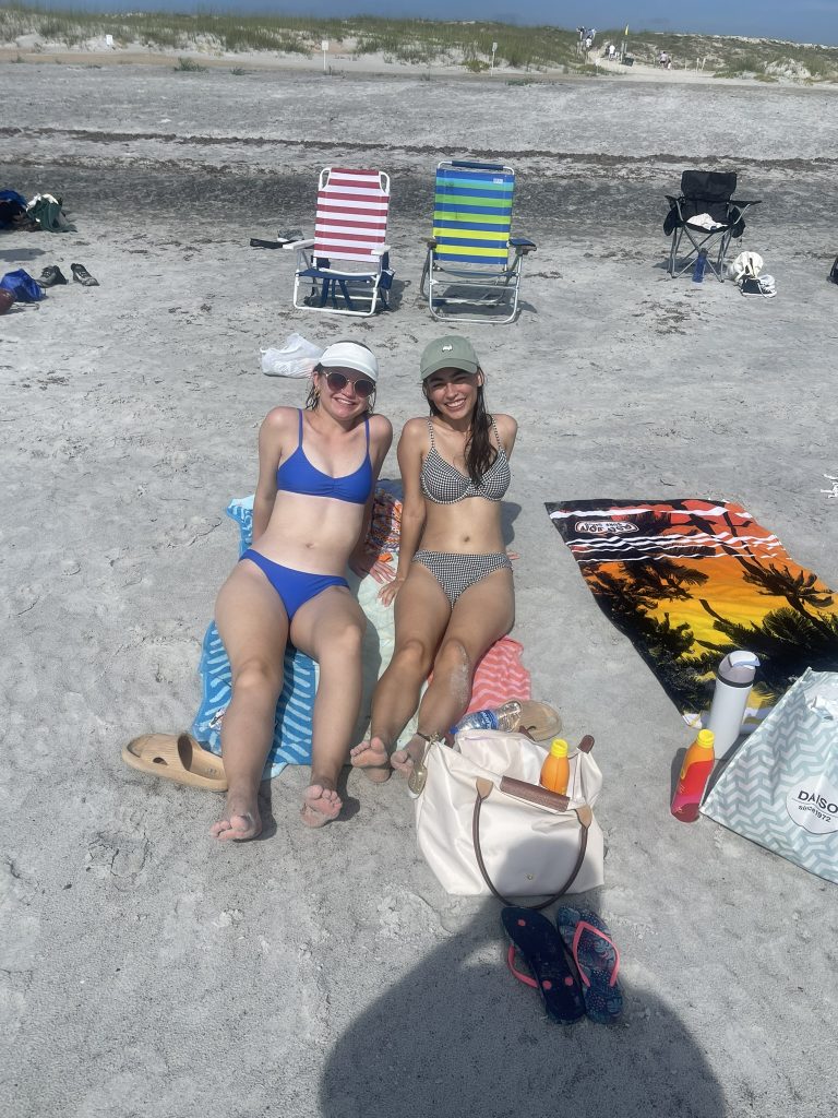 Two female students in their bathing suits, sitting on towels,  smiling and posing on the beach. 