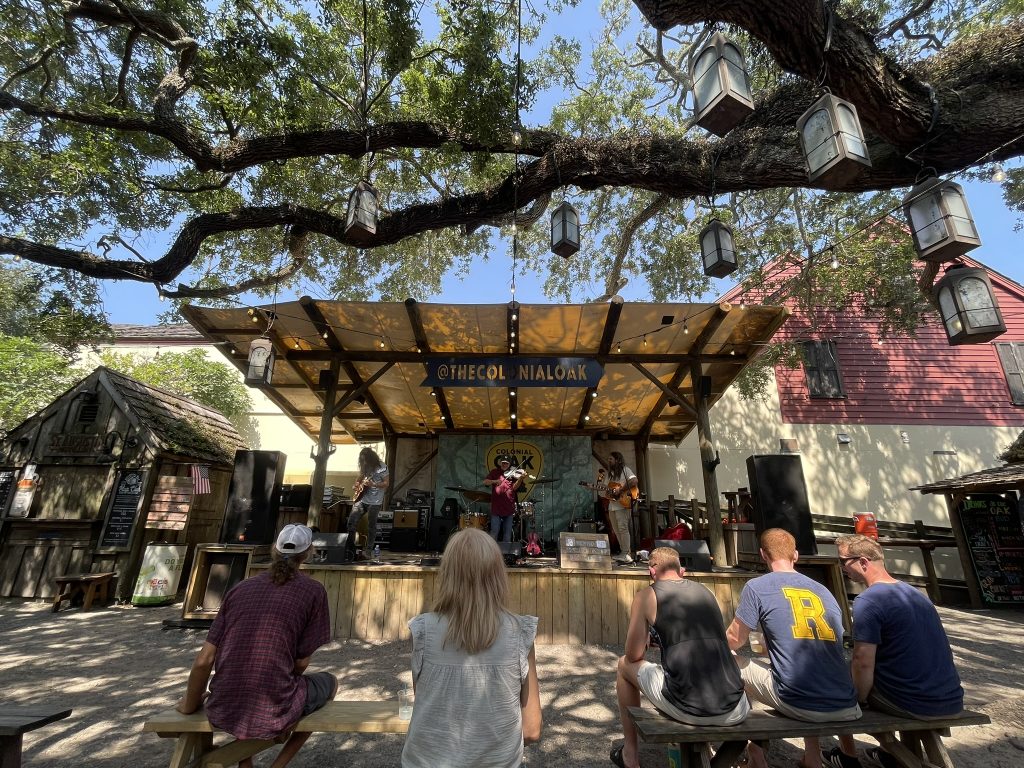 A band on stage, playing during a show, with it's audience sitting under the shade of a tree. 