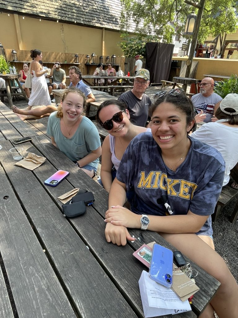 A trio of female student sitting at a picnic table and smiling. Other patrons are sitting behind them. 