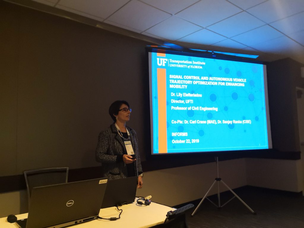 Dr. Elefteriadou stands to the left of a screen where the first slide of her presentation is projected. The presentation slide has the UF Transportation Institute Logo and reads: Signal Control and Autonomous Vehicle Trajectory Optimization for Enhancing Mobility. Dr. Lily Elefteriadou, Director, UFTI; Professor of Civil Engineering. Co-Pis: Dr. Carl Crane (MAE), Dr. Sanjay Ranka (CISE). INFORMS, October 22, 2019